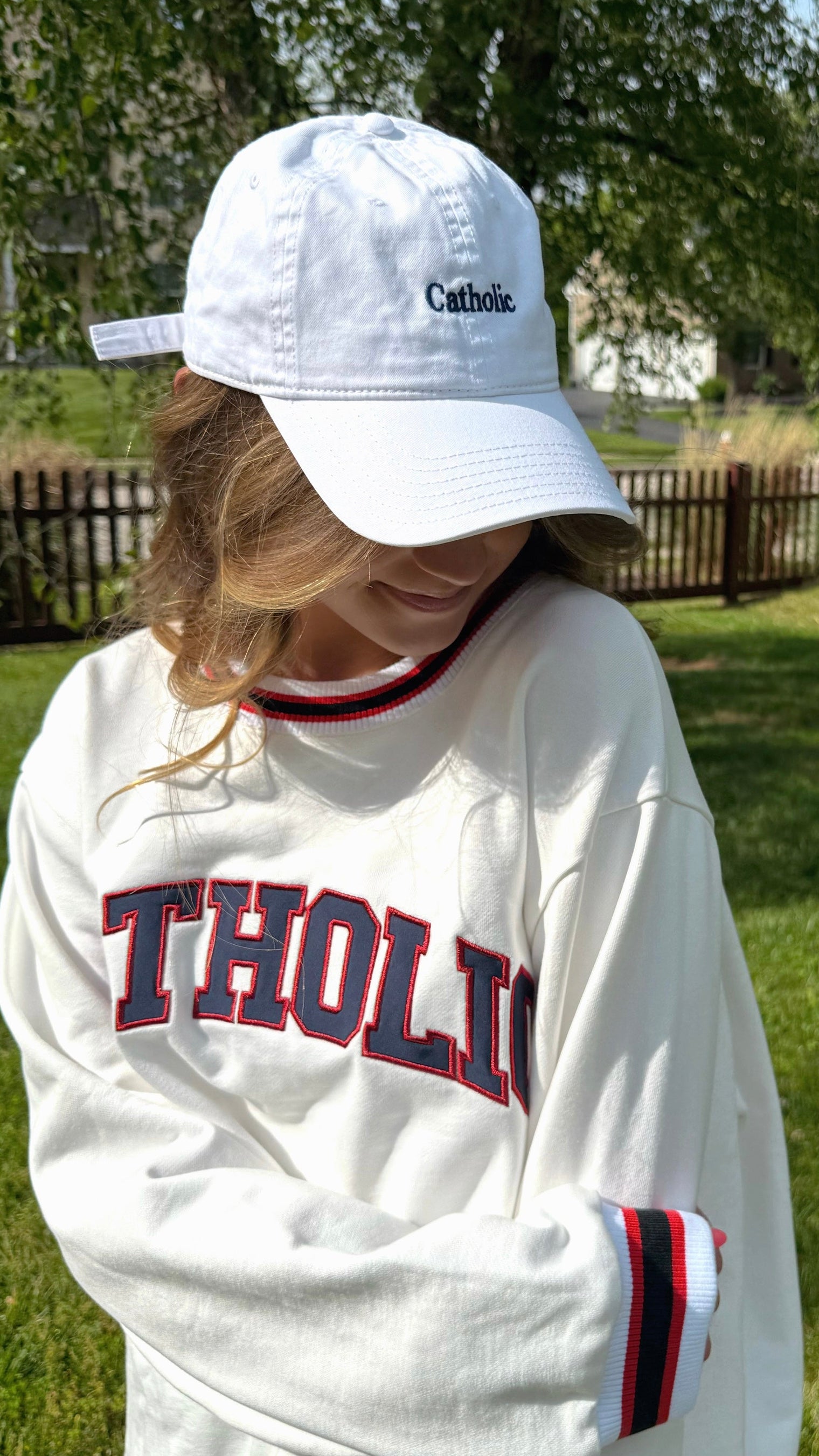 Person wearing a white Catholic cap and sweater with 'CATHOLIC' text outdoors.